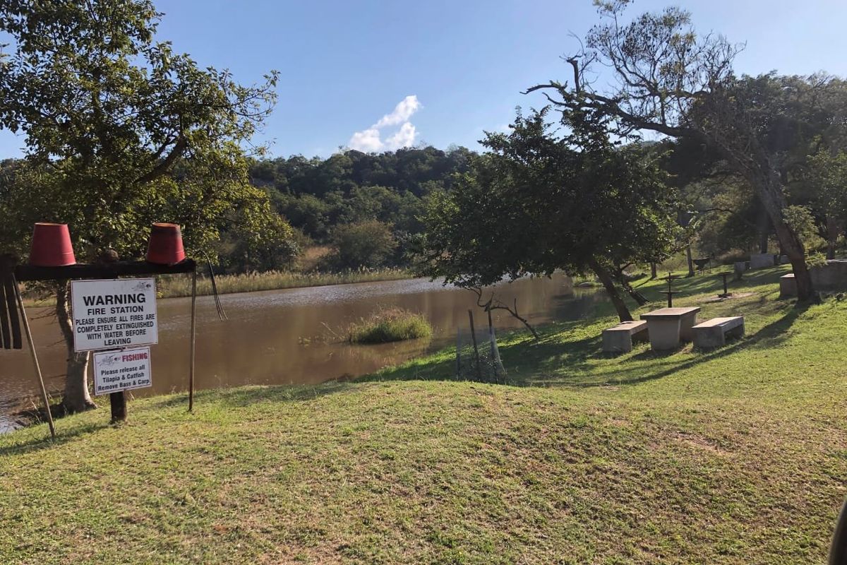 A scenic view of a lakefront area featuring established trees, grassy banks, and concrete picnic seating, ideal for outdoor recreation.