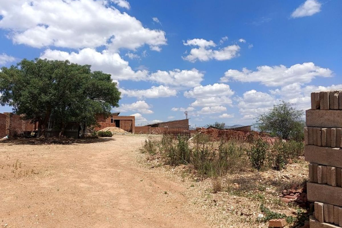 A vacant residential plot featuring an unpaved driveway, mature trees, and partial brick boundary walls, suitable for future development.