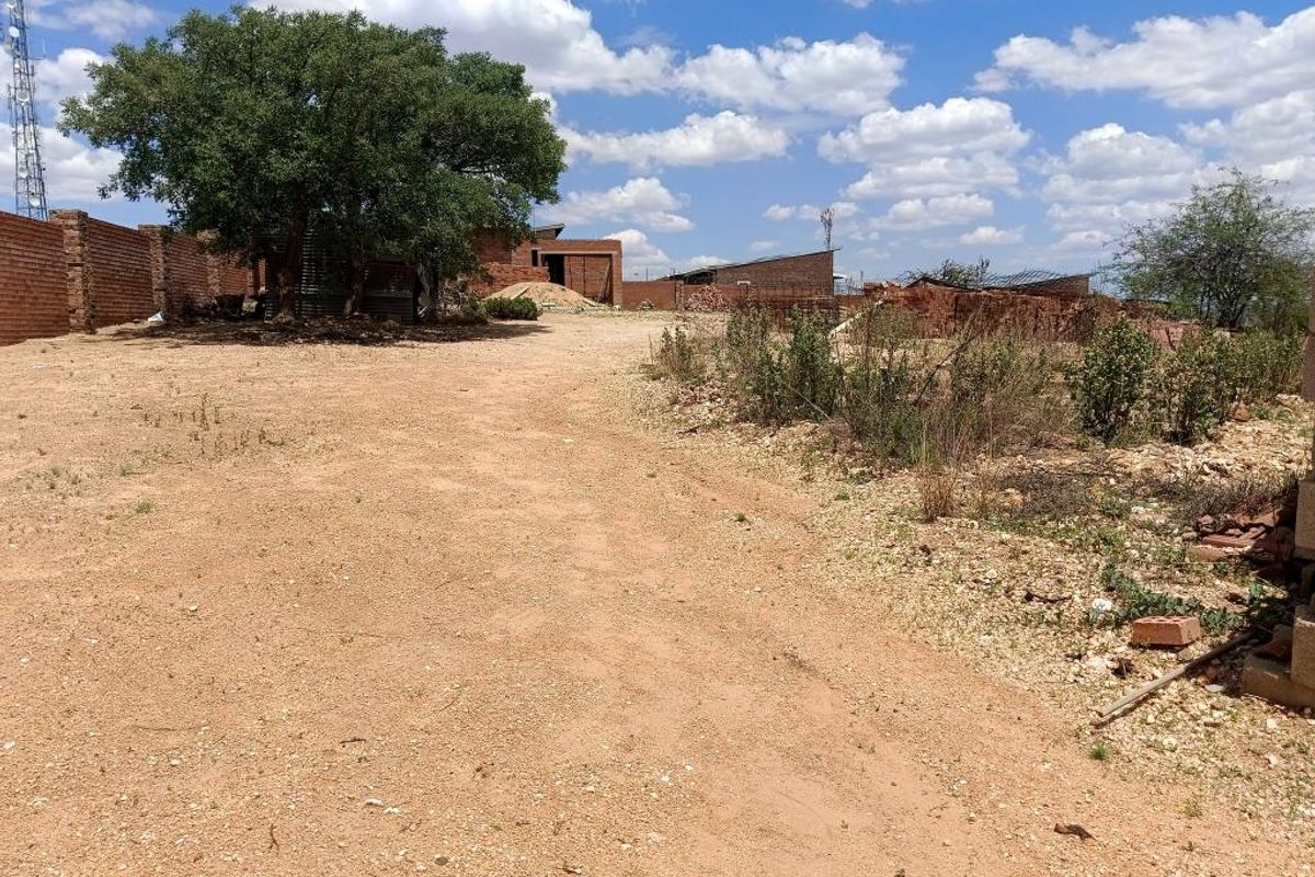 A view of a vacant residential plot featuring an unpaved driveway, a mature tree, and existing brick boundary walls.
