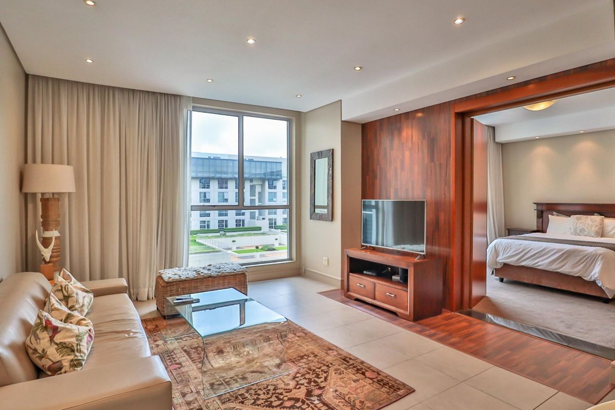 A well-maintained living room featuring tiled floors, recessed lighting, and a large window, with an open transition into the adjacent bedroom area.