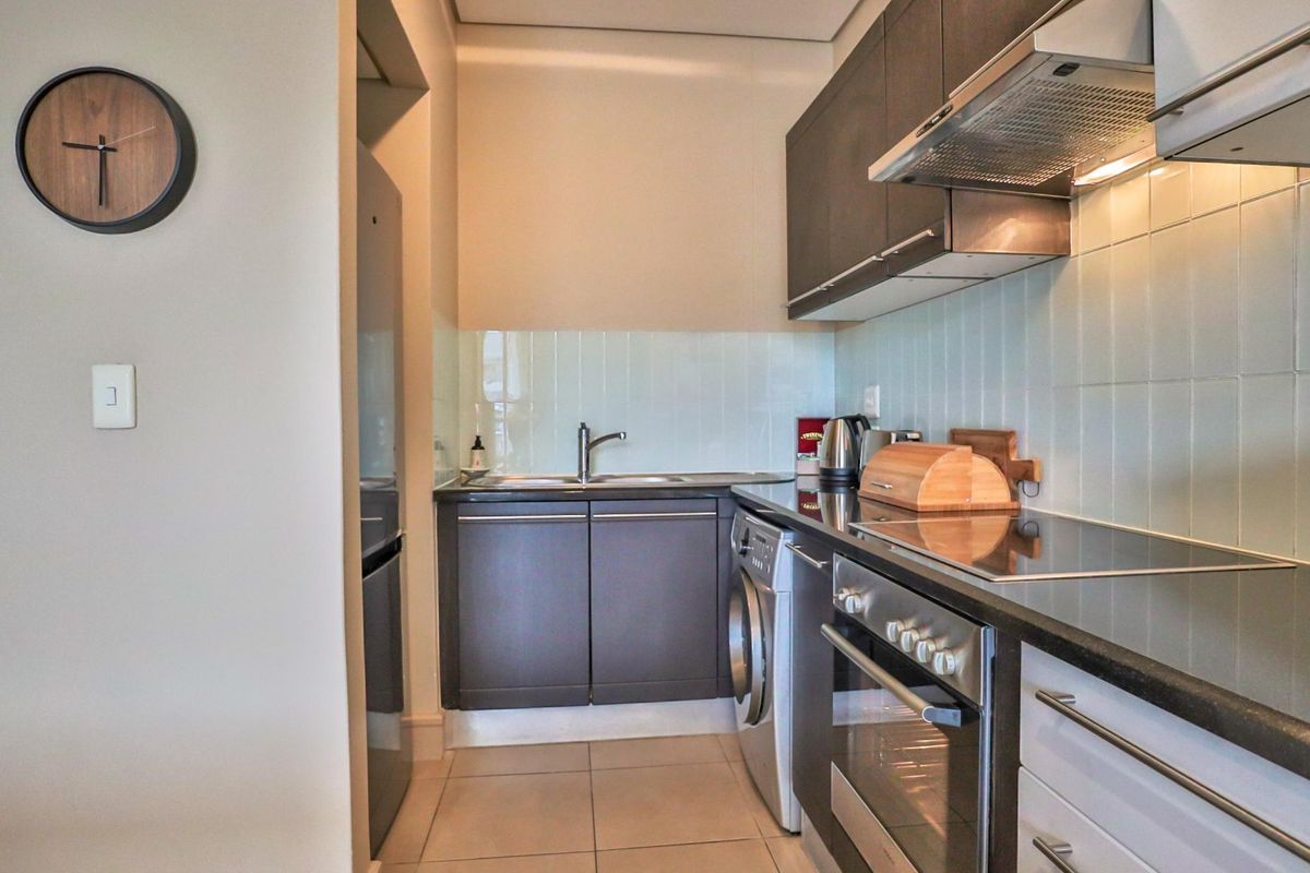 Modern compact kitchen featuring dark cabinetry, integrated stainless steel appliances, electric hob, extractor fan, and tiled backsplash with neutral floor tiles.