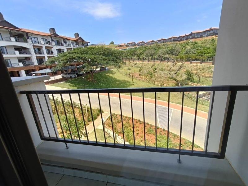 A balcony with a metal balustrade overlooking the apartment complex grounds, landscaped gardens, and surrounding hillside.