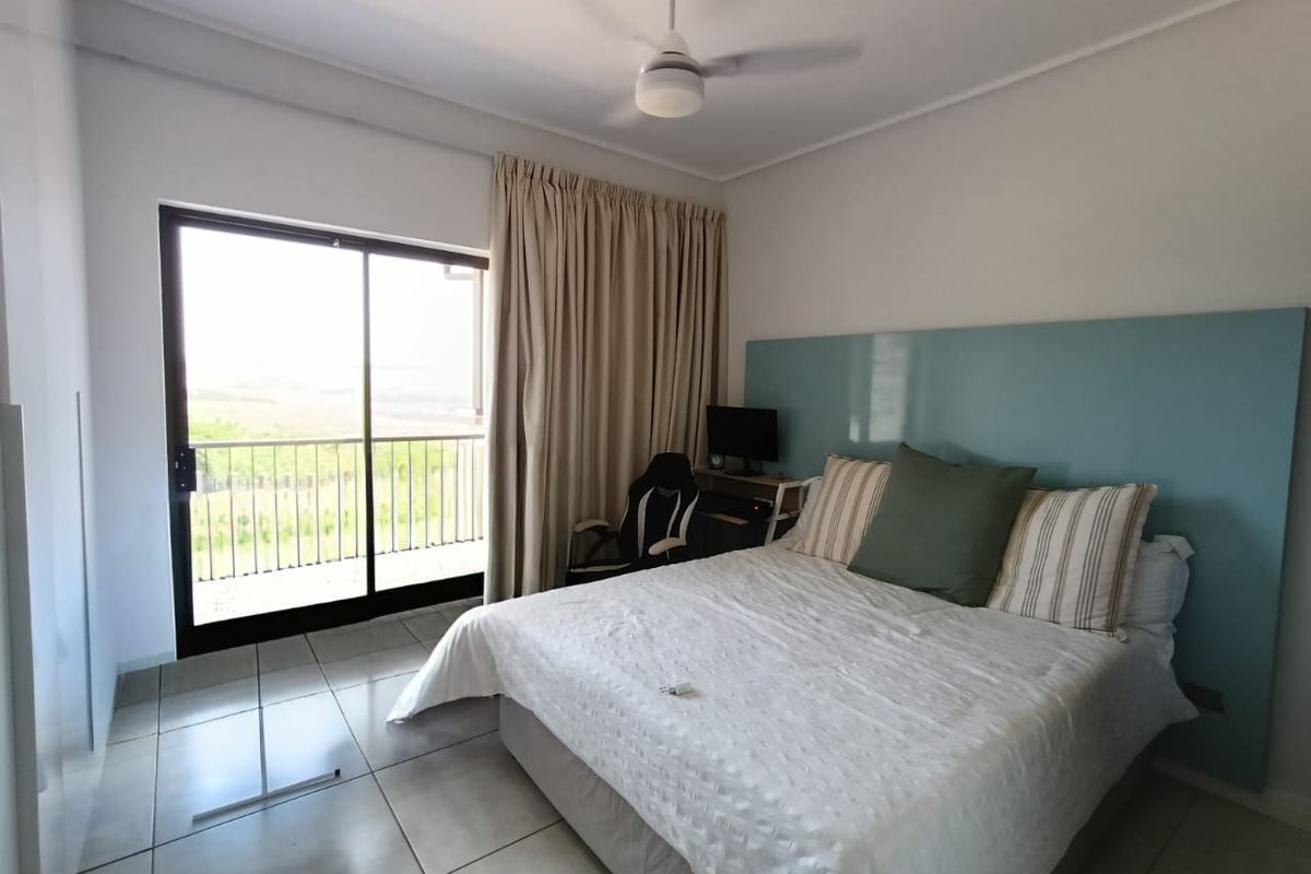 A bright bedroom featuring tiled flooring, a ceiling fan, and a large sliding glass door providing access to a balcony with scenic views.