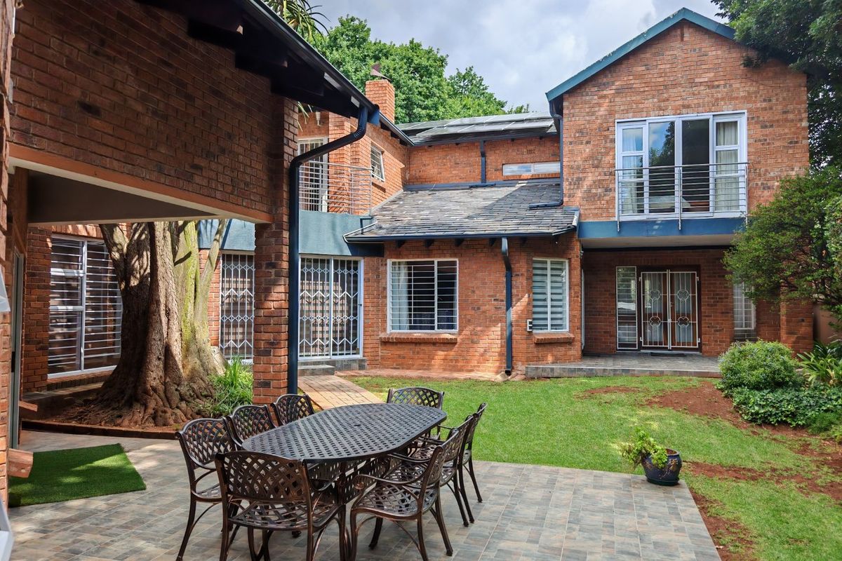 A spacious rear exterior view featuring a brick facade, a covered patio area, a balcony, and a well-maintained garden.