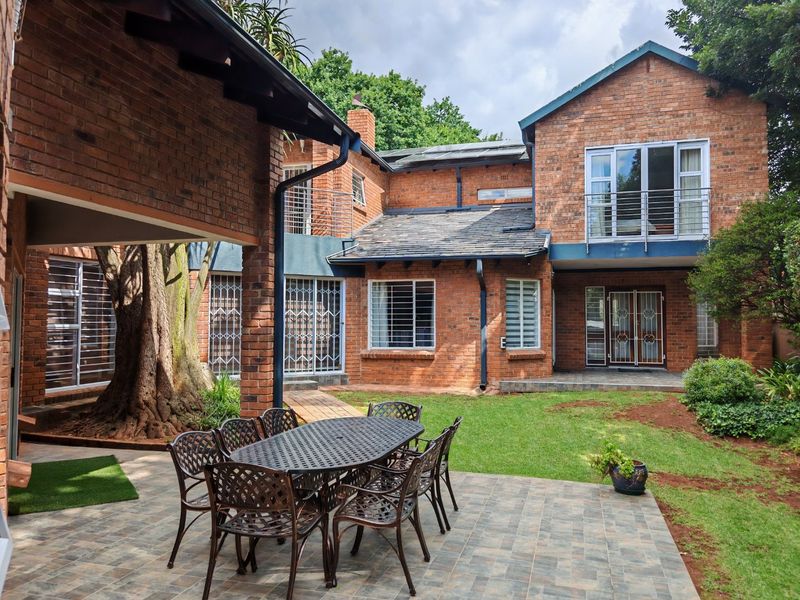 A spacious rear exterior view featuring a brick facade, a covered patio area, a balcony, and a well-maintained garden.