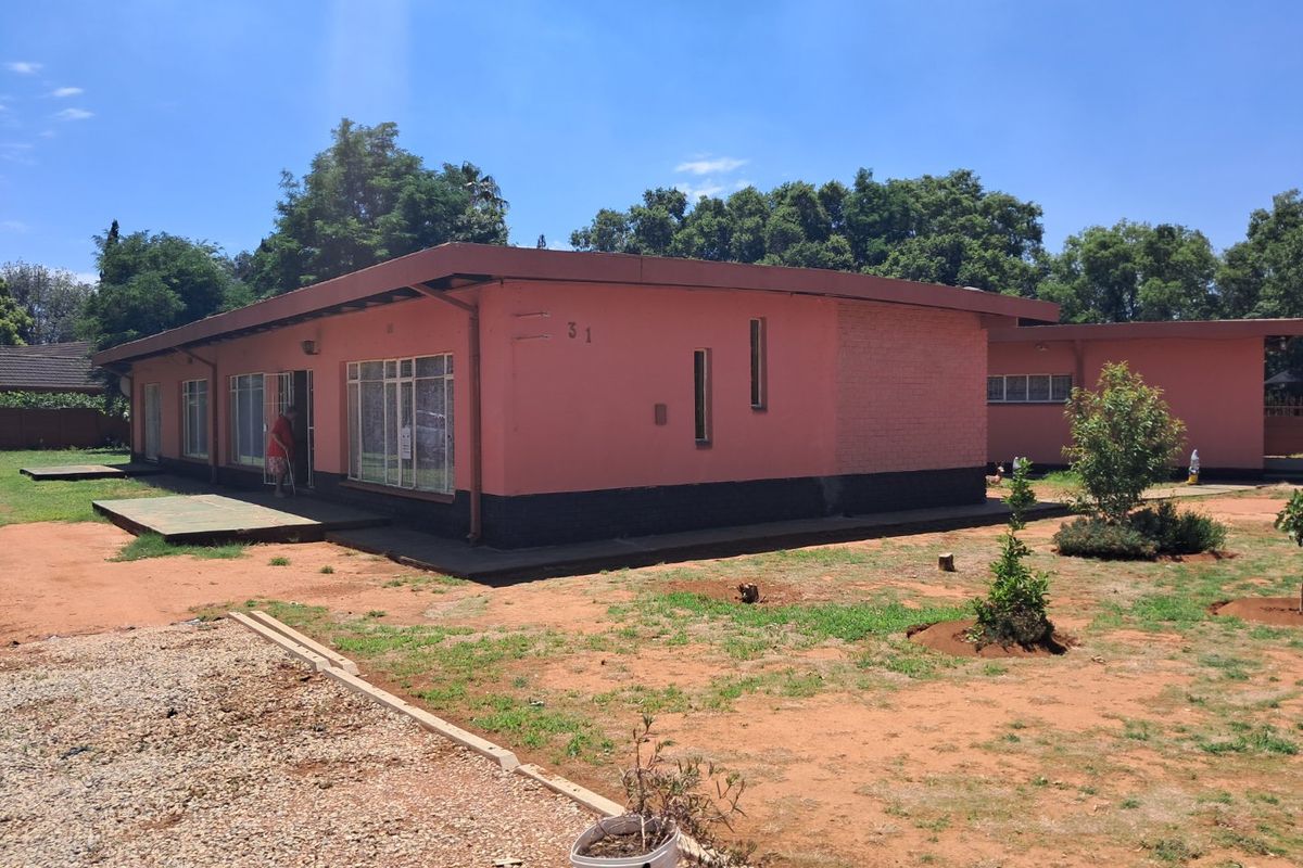 Rear view of a single-story residential property featuring brick construction, metal window frames, and a concrete patio area set within a large garden.