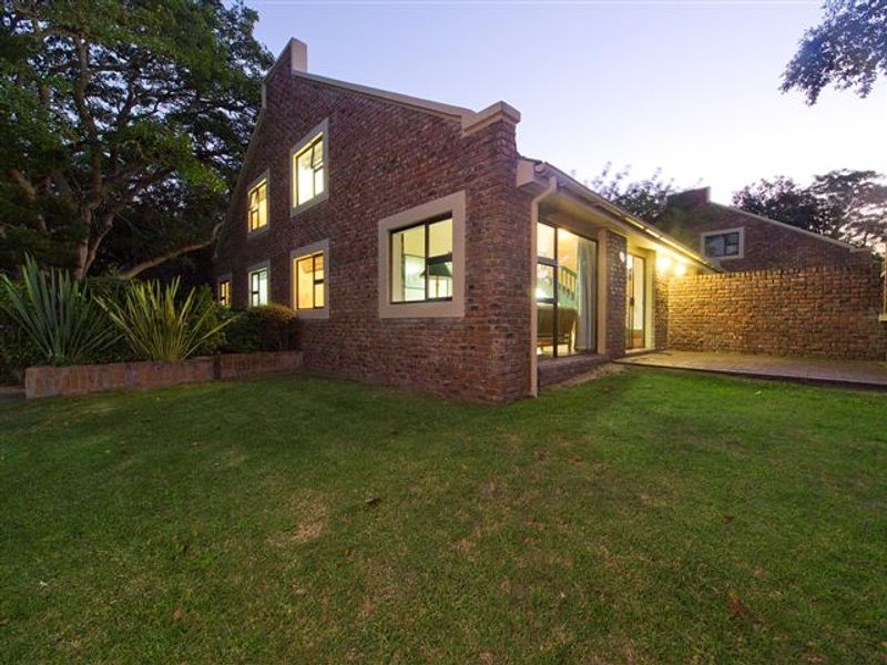 A well-maintained rear exterior view featuring an exposed brick facade, paved patio area, and a neatly landscaped lawn with mature trees.