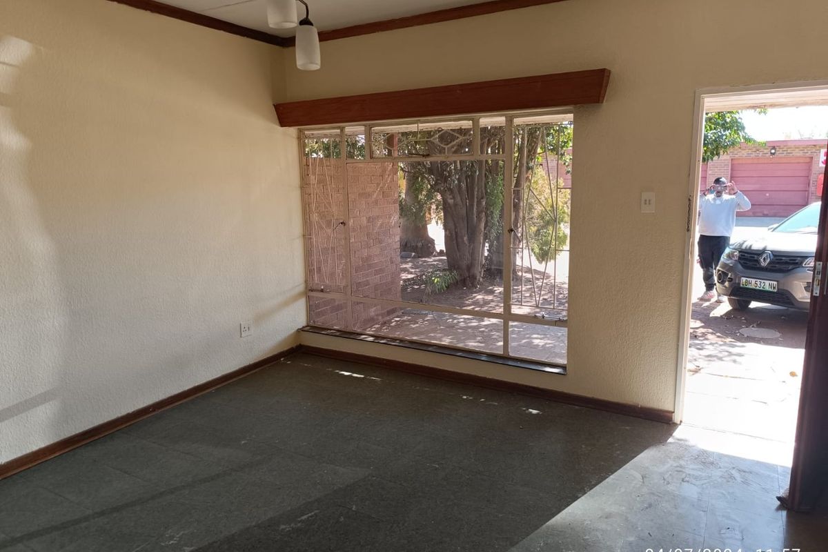 An empty living room featuring large metal-framed windows with security bars, tiled flooring, and a simple pendant light fixture.