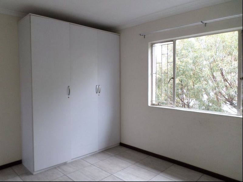 A clean, empty bedroom featuring neutral tiled flooring, a built-in white wardrobe, and a window fitted with security bars.