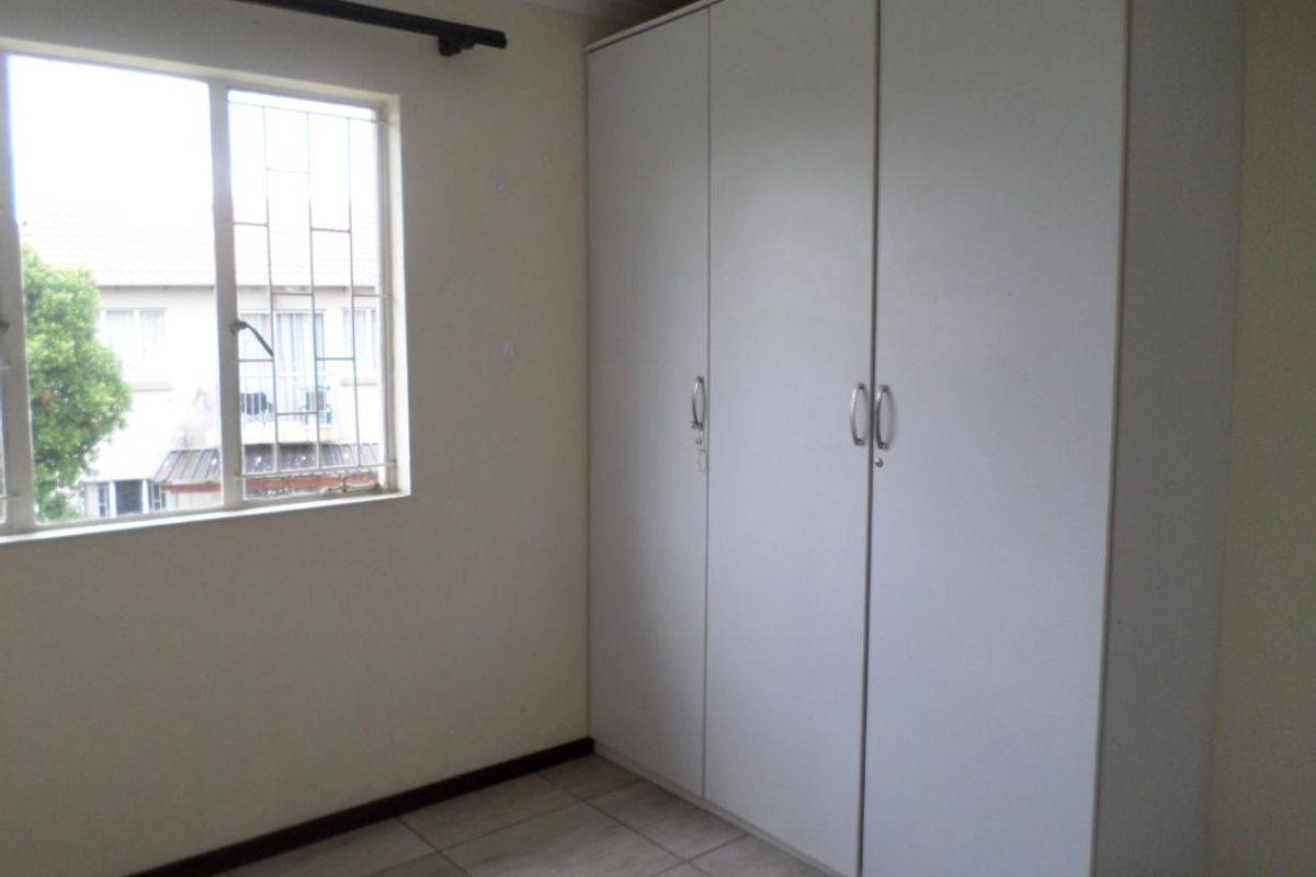 A clean, empty bedroom featuring neutral tiled flooring, a large built-in wardrobe, and a window fitted with security bars.
