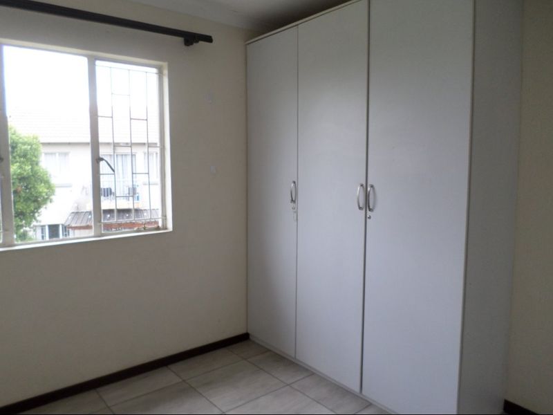 A clean, empty bedroom featuring neutral tiled flooring, a large built-in wardrobe, and a window fitted with security bars.