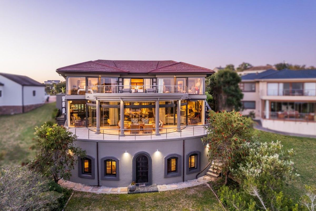 A multi-level luxury residence featuring a curved rear facade, expansive glass sliding doors, a spacious balcony with stainless steel balustrades, and manicured garden surroundings.