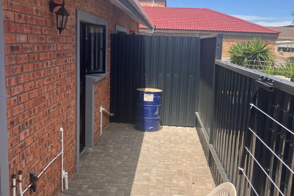 A paved rear exterior area featuring a brick facade, metal privacy screening, and external plumbing fixtures, suitable for a utility or service space.