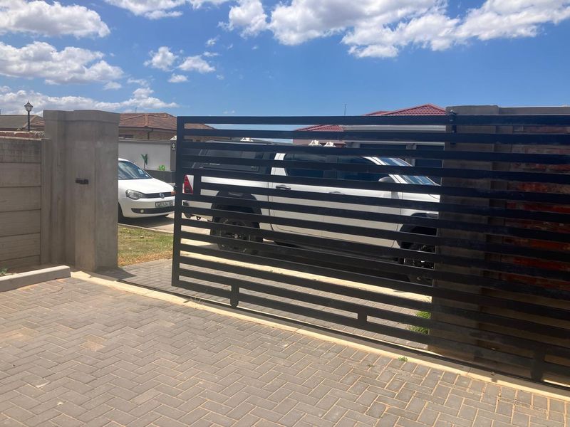 A modern horizontal slat sliding gate secures the front entrance, featuring a paved driveway and concrete boundary walling.