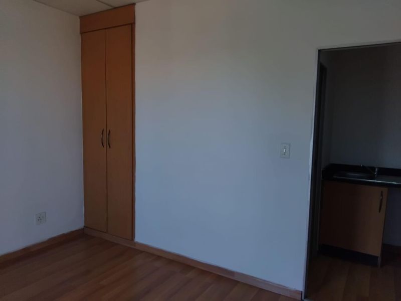 A clean, empty bedroom featuring laminate flooring and a built-in wooden wardrobe, with a view into an adjacent kitchenette area.