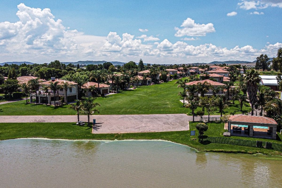 Aerial view of a residential estate featuring a lake, expansive green lawns, paved driveways, and a communal pool area with a covered patio.
