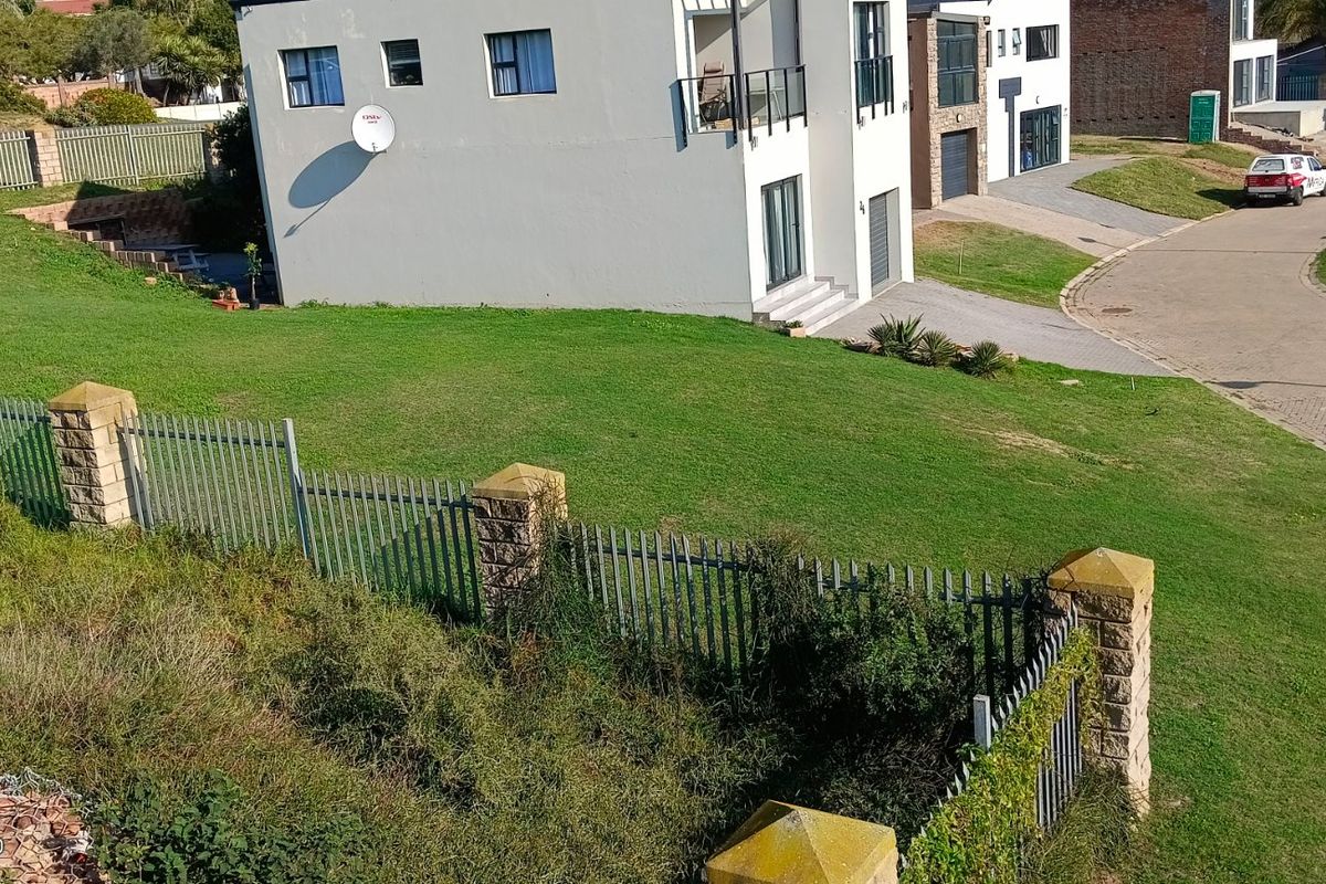 An elevated view of a vacant residential plot featuring established lawn, perimeter fencing with stone pillars, and adjacent paved street access.