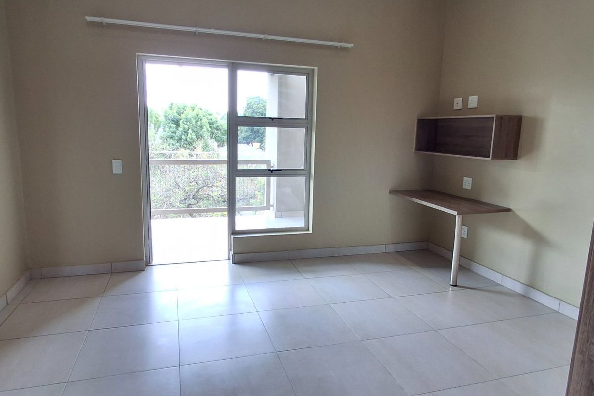 A bright, empty bedroom featuring tiled floors, a built-in desk with shelving, and a sliding glass door leading to a private balcony.