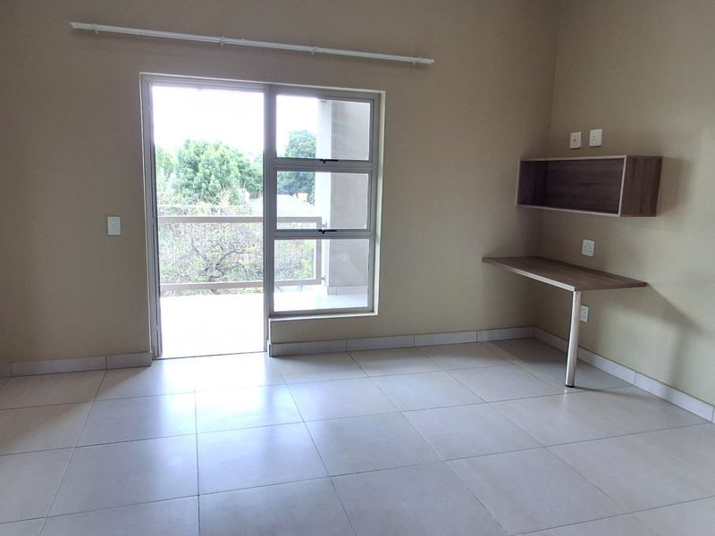 A bright, empty bedroom featuring tiled floors, a built-in desk with shelving, and a sliding glass door leading to a private balcony.
