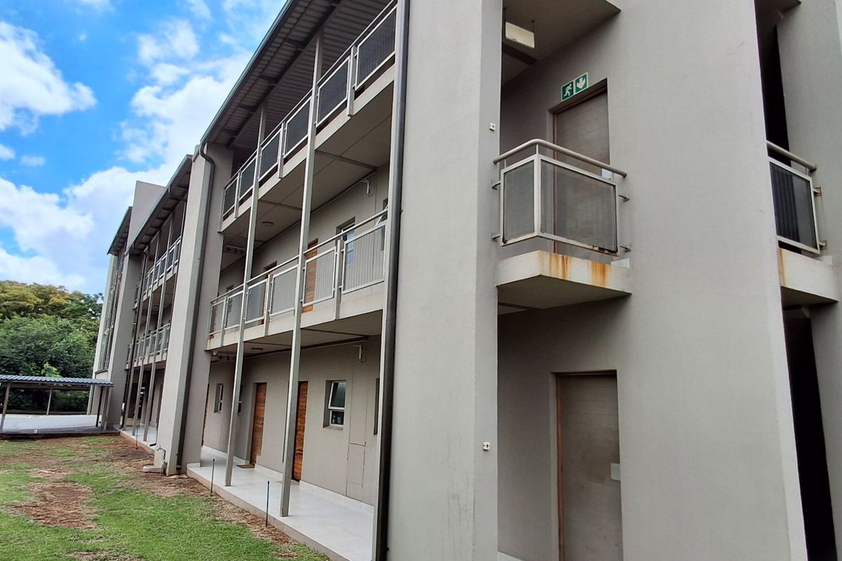 Exterior view of a multi-story apartment building featuring concrete walkways, metal balcony railings, and an adjacent carport area with surrounding lawn.