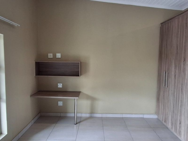 A clean, empty bedroom featuring neutral tiled flooring, ample built-in wooden cupboards, and a convenient wall-mounted desk and shelf unit.