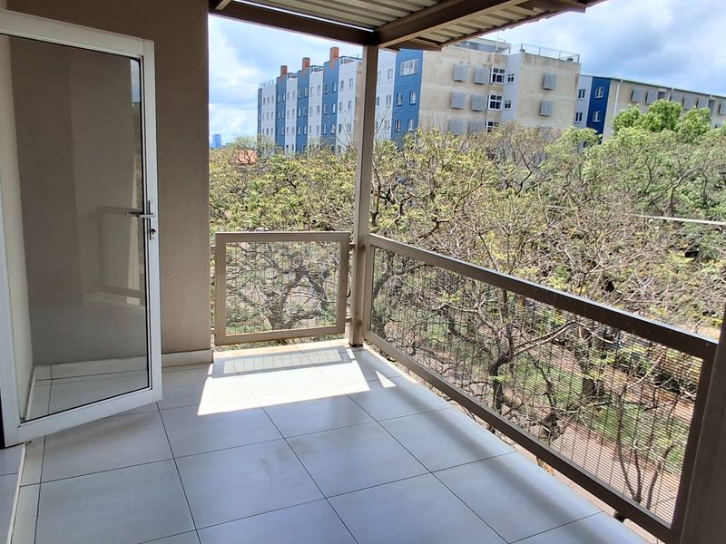 A clean, covered balcony featuring tiled flooring and a metal mesh railing, offering an open outdoor space accessible via a sliding glass door.