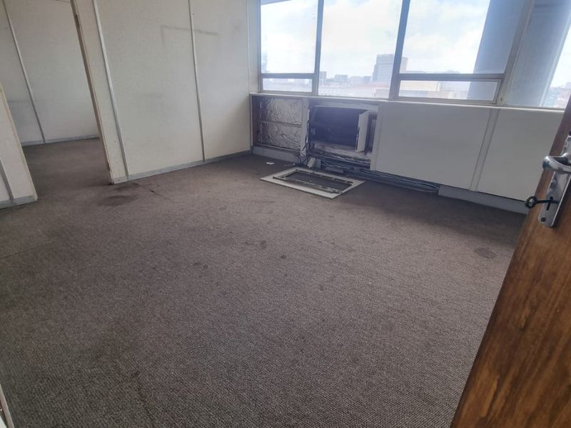 Empty office space featuring large windows, carpet flooring, and suspended ceiling, currently requiring maintenance and cosmetic updates to the wall panels and HVAC unit.