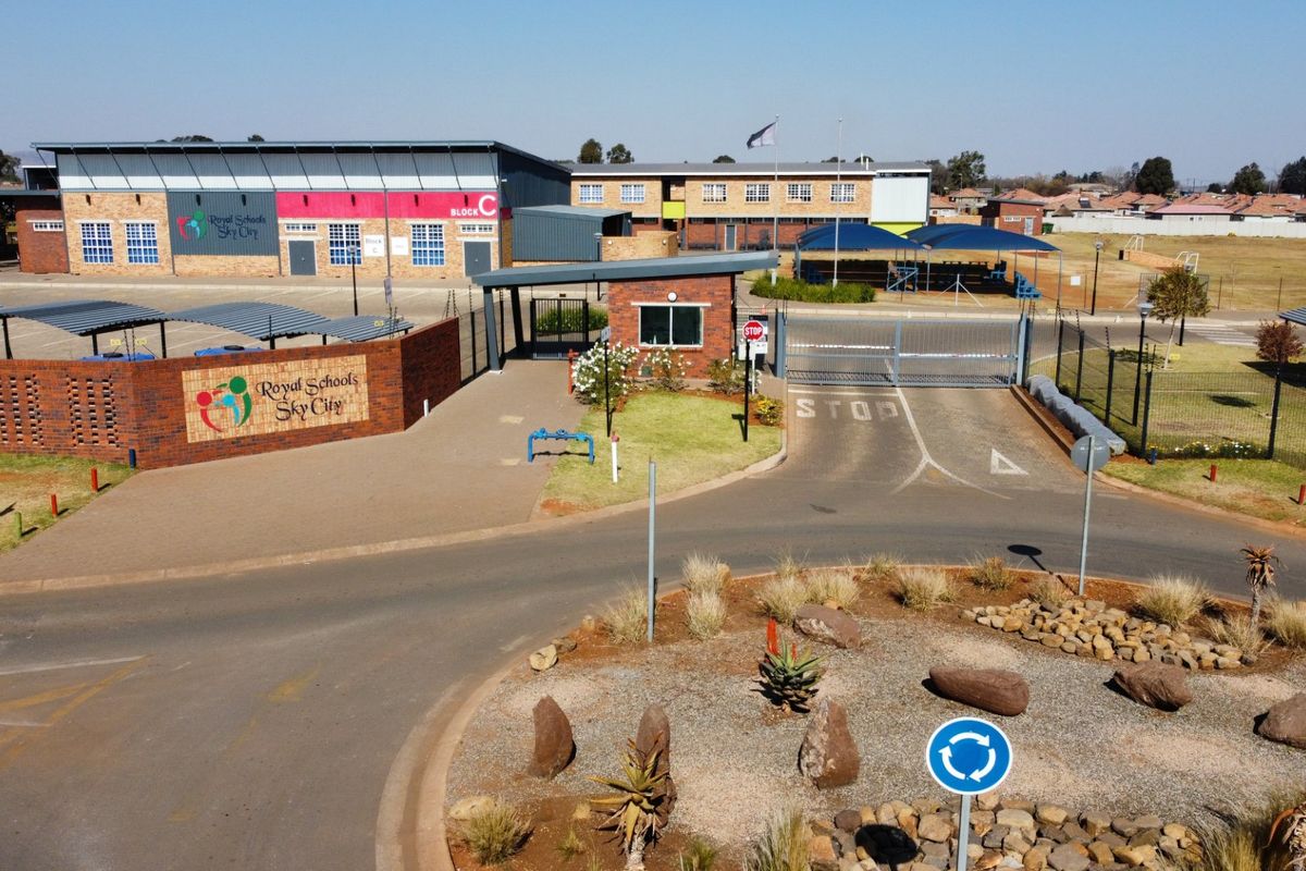 Aerial view of the school entrance featuring a secure gated access point, brick boundary wall, paved driveway, and covered parking areas.