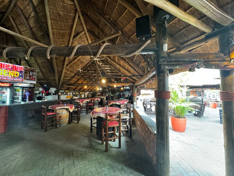 A rustic, open-air bar area featuring a traditional thatched roof, exposed timber support beams, a built-in bar counter, and durable paved flooring.