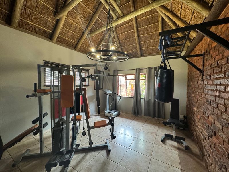 A dedicated home gym space featuring an exposed thatch roof, rustic brick feature wall, tiled flooring, and a decorative wrought iron chandelier.