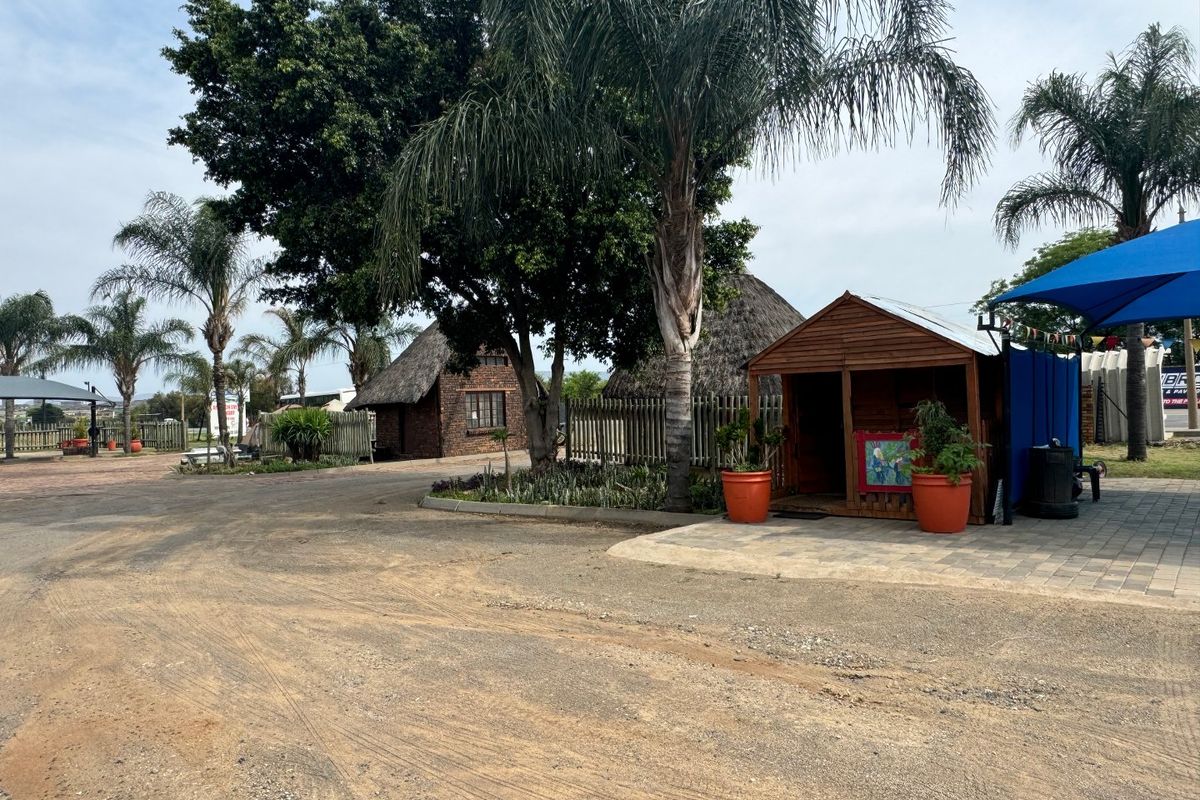 Outdoor commercial area featuring thatched roof structures, a paved driveway, and a timber shed under a shade sail.