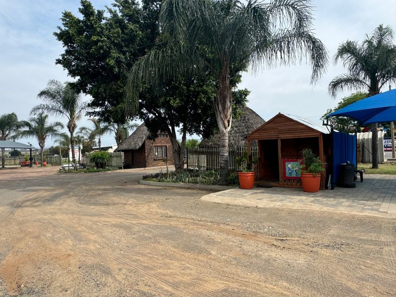 Outdoor commercial area featuring thatched roof structures, a paved driveway, and a timber shed under a shade sail.