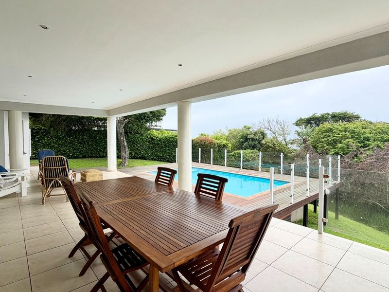 Spacious covered patio featuring tiled flooring and recessed lighting, overlooking a swimming pool with a glass balustrade and lush garden views.