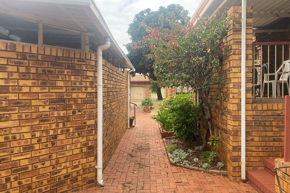 A paved walkway between two face brick townhouse structures, featuring established garden beds and access to a covered patio area.