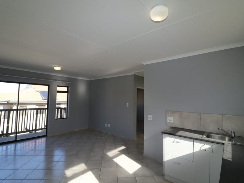 An open-plan living and kitchen area featuring tiled flooring, neutral grey walls, and a sliding door leading to a balcony.