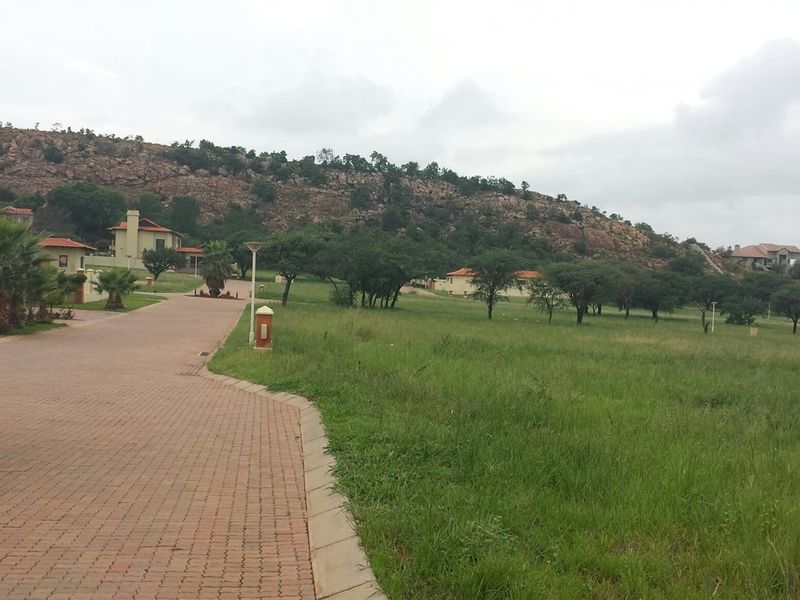 A view of a residential street featuring a paved driveway, manicured verge, and established trees set against a natural rocky hillside backdrop.