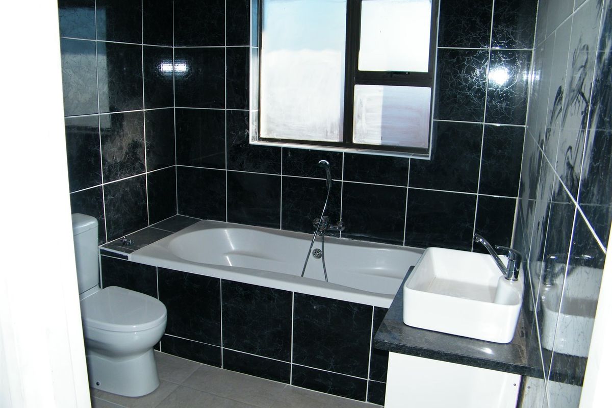 A bathroom featuring black marble-effect wall tiles, a built-in bathtub with shower mixer, a white ceramic basin, and a toilet.