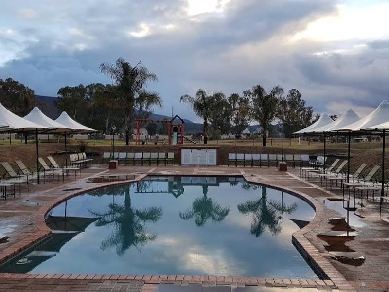 A well-maintained outdoor swimming pool area featuring a paved deck, multiple tensile shade structures, and surrounding landscaped grounds.