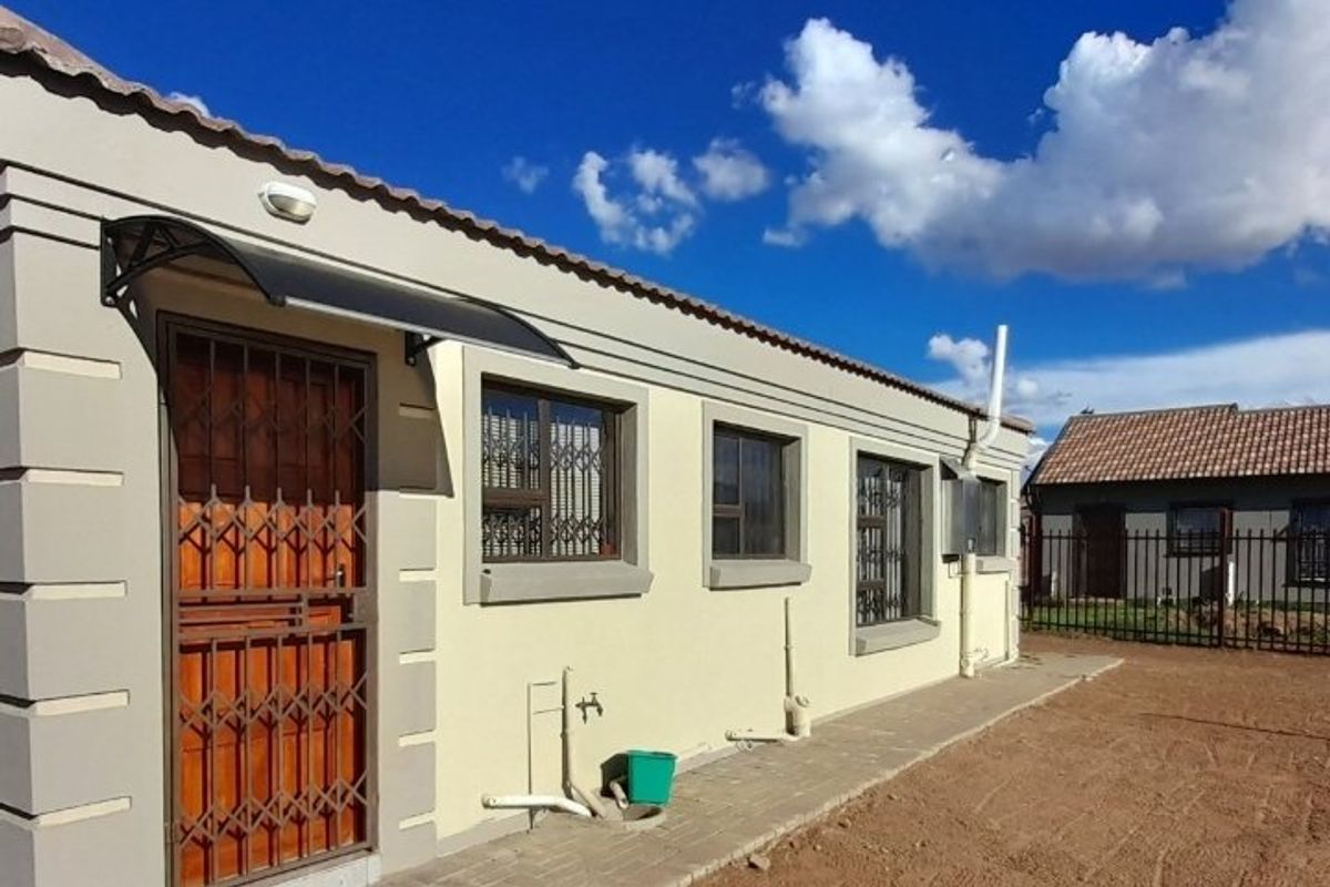 Rear exterior view of the house featuring a paved walkway, security gate, and external plumbing fixtures against a clean rendered facade.