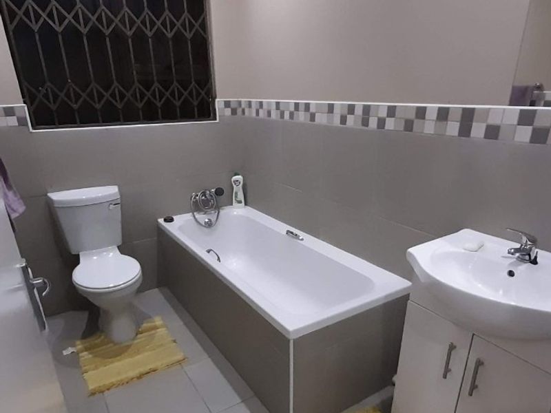 A functional bathroom featuring a built-in bathtub, a vanity cabinet with a basin, and a toilet, finished with neutral wall and floor tiles.