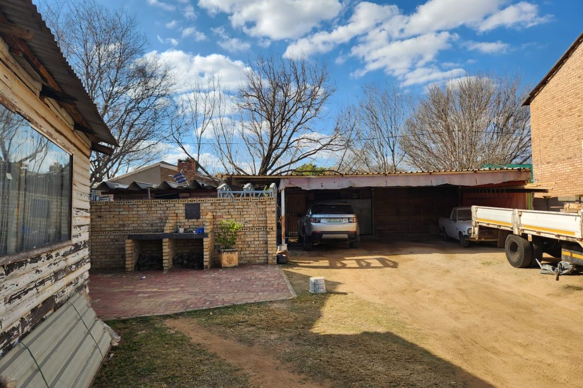 Rear exterior view featuring a brick built-in braai area, covered parking structure, and unpaved yard space with weathered building finishes.