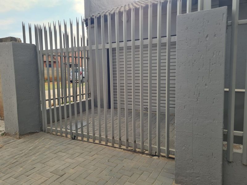 Front exterior view featuring a secure steel gate, roller shutter garage door, and paved driveway area.