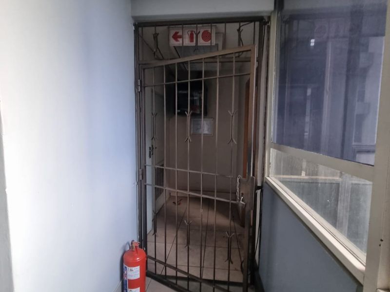 A commercial hallway featuring tiled flooring, a security gate, and emergency safety equipment, showing signs of wear on the ceiling and walls.