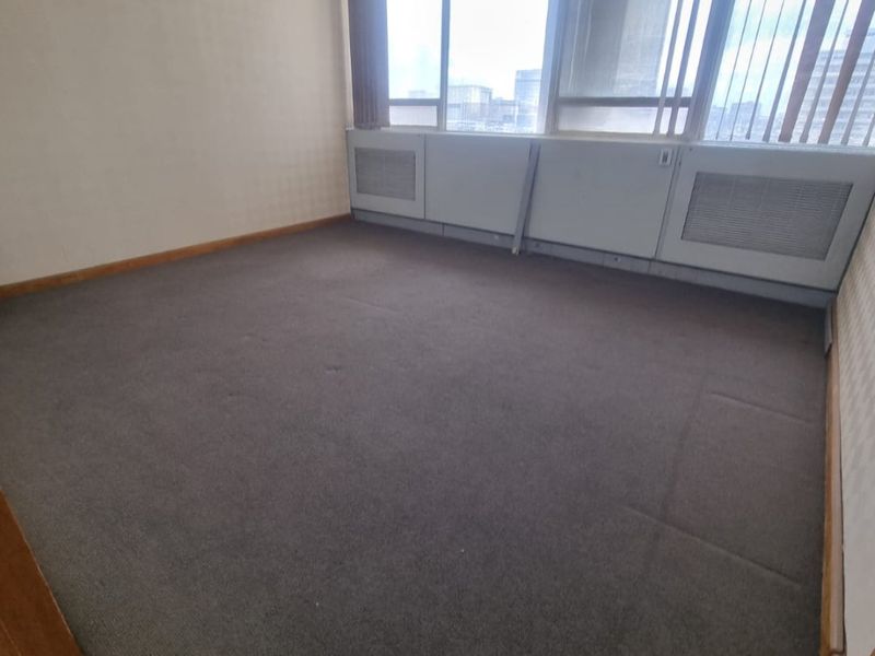 An empty commercial office space featuring large windows with vertical blinds, built-in HVAC units, a suspended ceiling, and carpeted flooring.