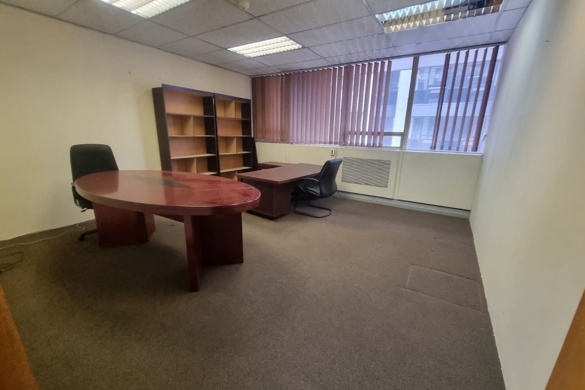 A functional office space featuring a suspended ceiling, recessed lighting, vertical blinds, and carpeted flooring, suitable for a professional work environment.