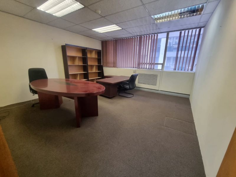 A functional office space featuring a suspended ceiling, recessed lighting, vertical blinds, and carpeted flooring, suitable for a professional work environment.