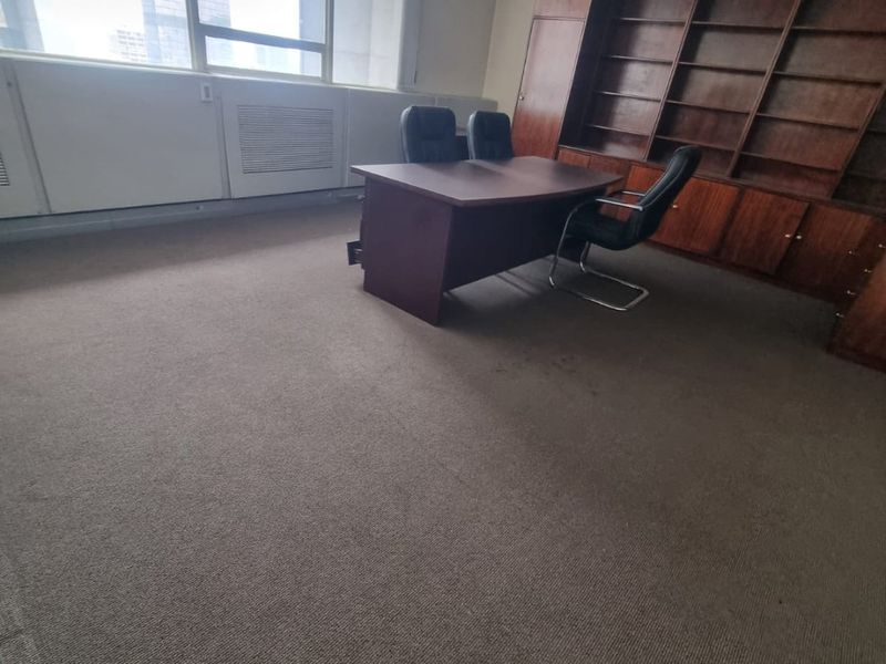 A commercial office space featuring built-in wooden shelving, large windows providing natural light, and carpeted flooring.
