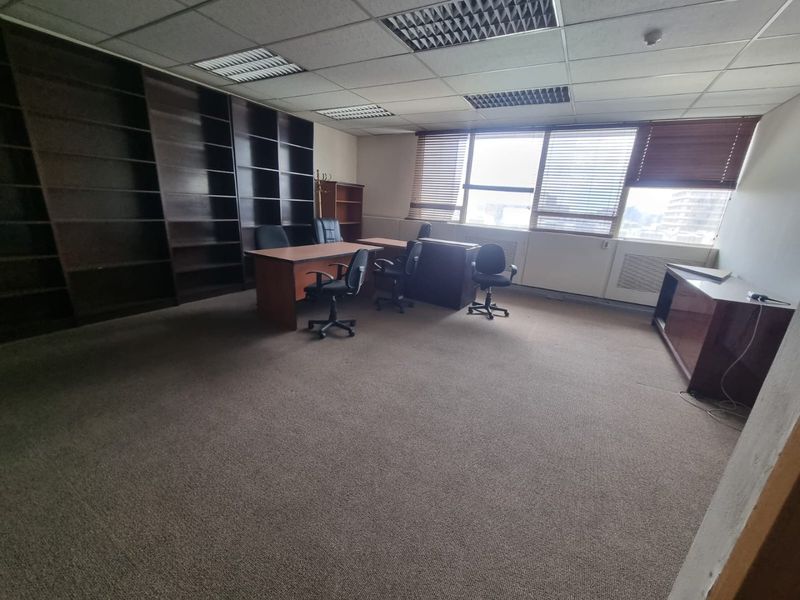 Spacious commercial office featuring built-in floor-to-ceiling shelving, suspended ceiling with recessed lighting, and large windows with venetian blinds.