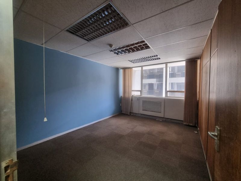 An empty office space featuring a suspended ceiling, built-in cabinetry, carpeted flooring, and large windows providing natural light.
