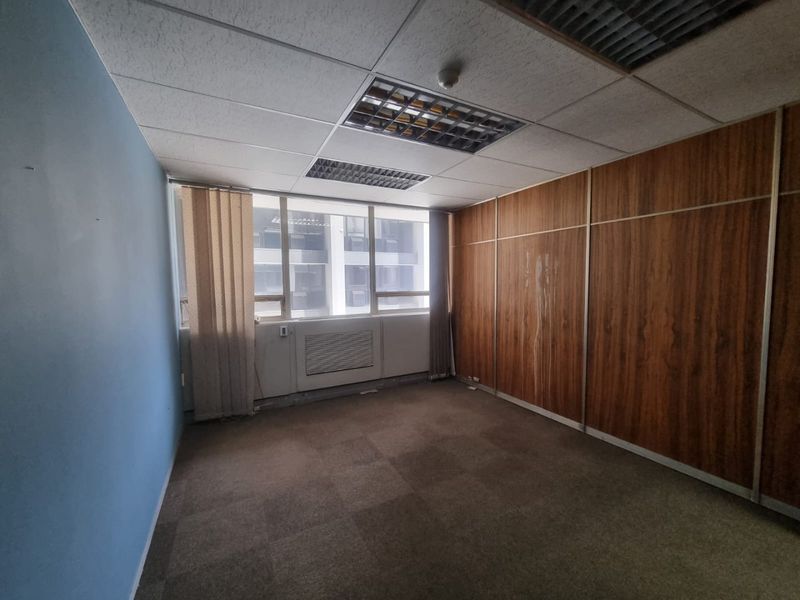 An empty commercial office space featuring wood veneer wall paneling, carpeted flooring, and recessed ceiling lighting with large windows providing natural light.